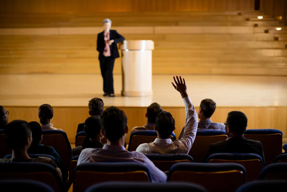 A woman in a black coat confidently standing at a podium and comunicating with her audience.